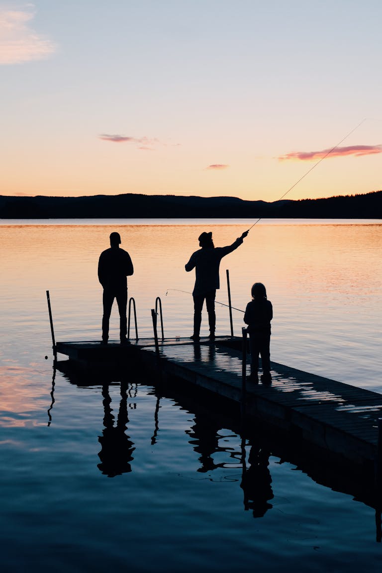 A serene scene of a family fishing together on a lake dock at sunset, creating silhouettes on the water.