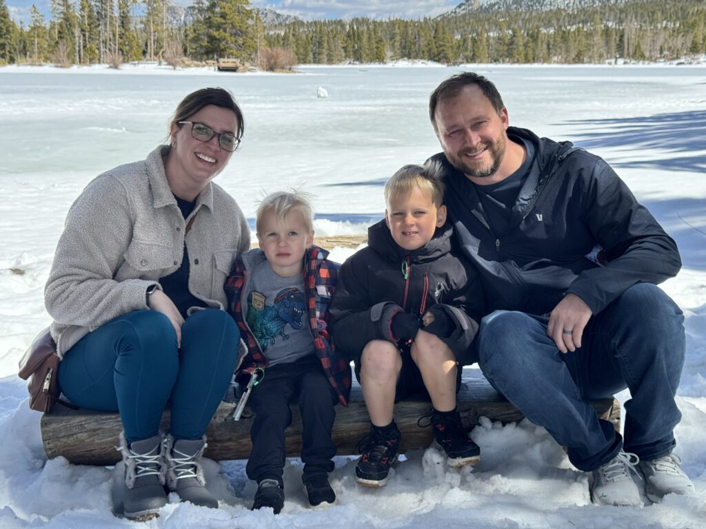 Family with mom, dad and two sons sitting on a log in front of a frozen lake surrounded by snow