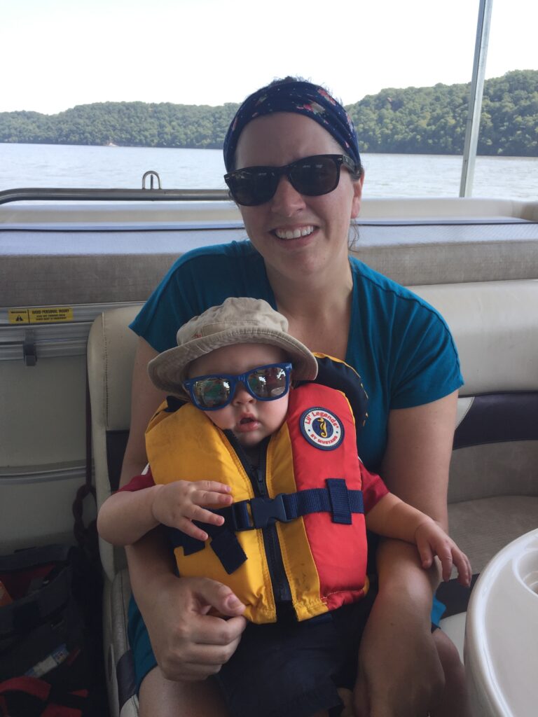 Mom with baby in a bucket hat, sunglasses and life jacket sitting on a dock at the lake