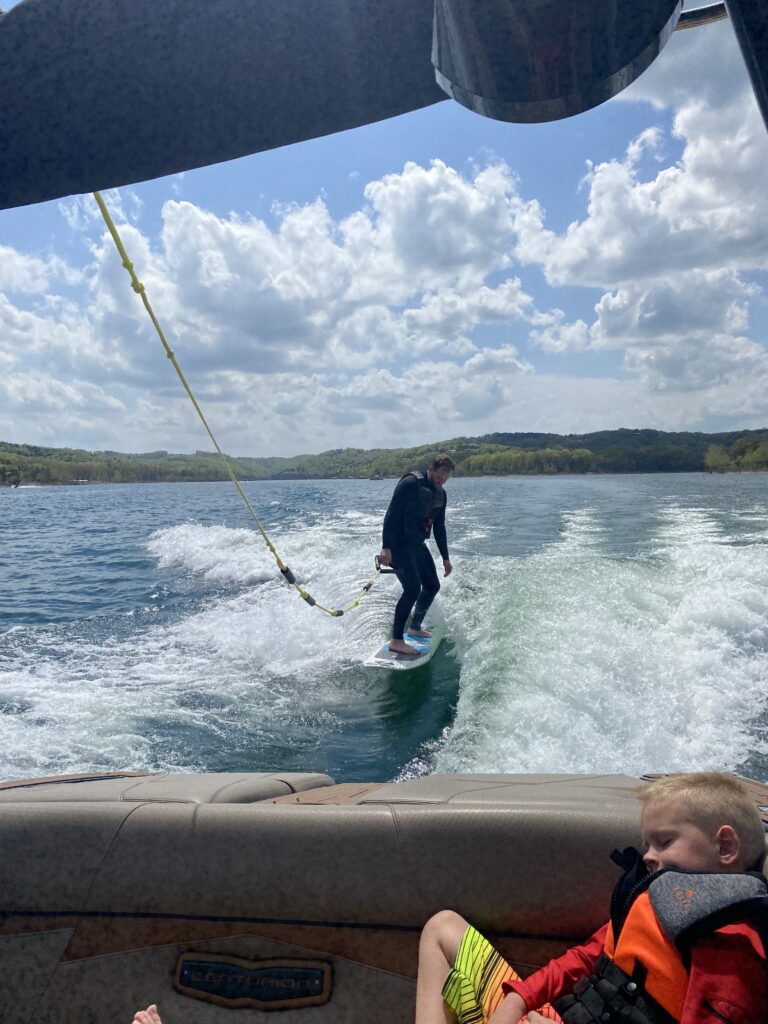 Dad wake surfing off the back of a boat with son sleeping in the back seat