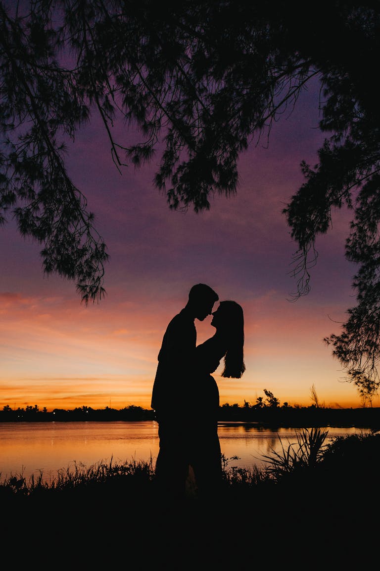 A couple embraces in a romantic silhouette by the lake during a vibrant sunset.