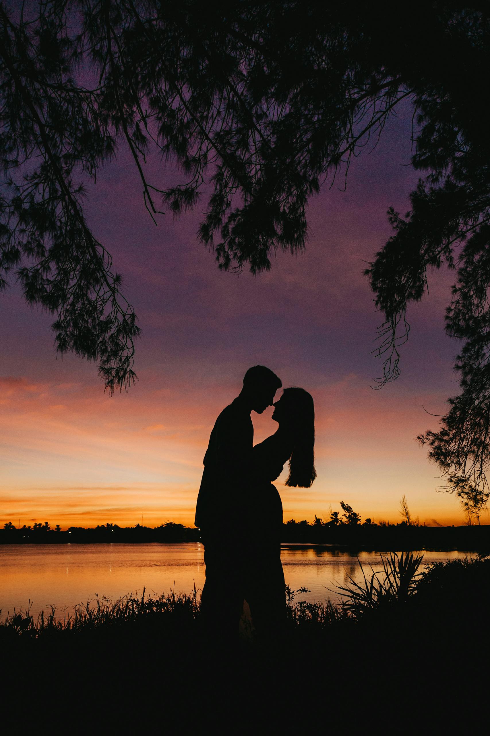 A couple embraces in a romantic silhouette by the lake during a vibrant sunset.