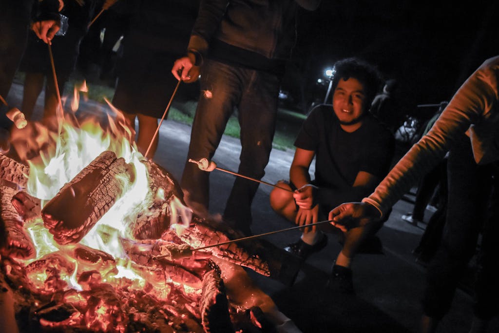 A group enjoys roasting marshmallows and socializing around a vibrant campfire at night.