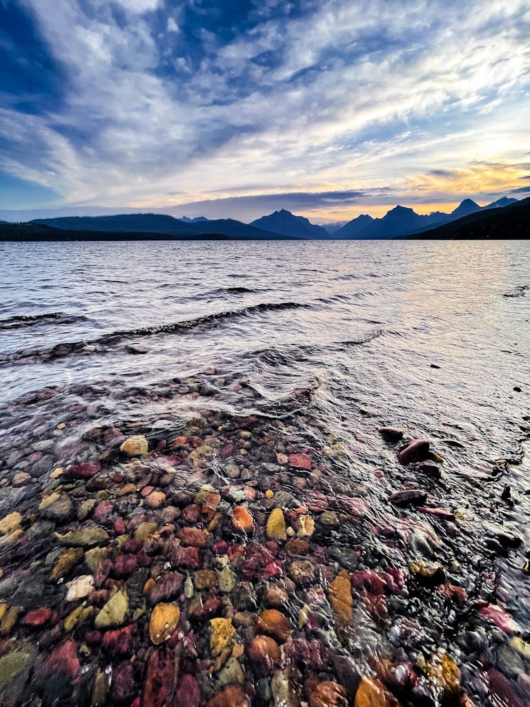 A tranquil view of Lake McDonald with colorful rocks and distant mountains at dusk.