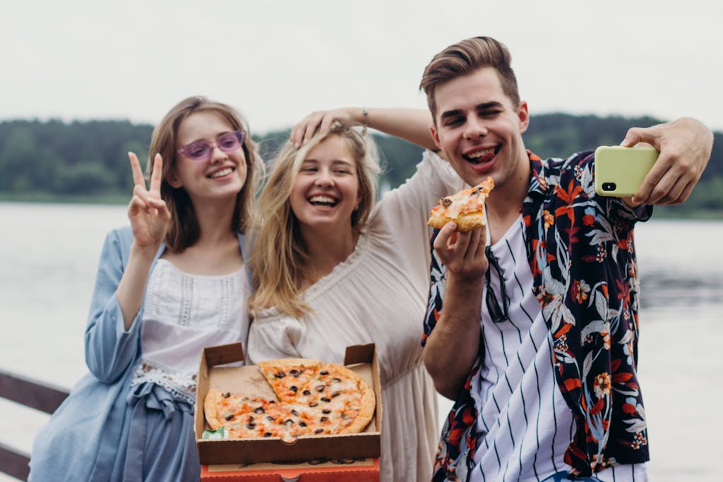 Group of friends enjoying pizza and taking a selfie by the lake, capturing fun moments.