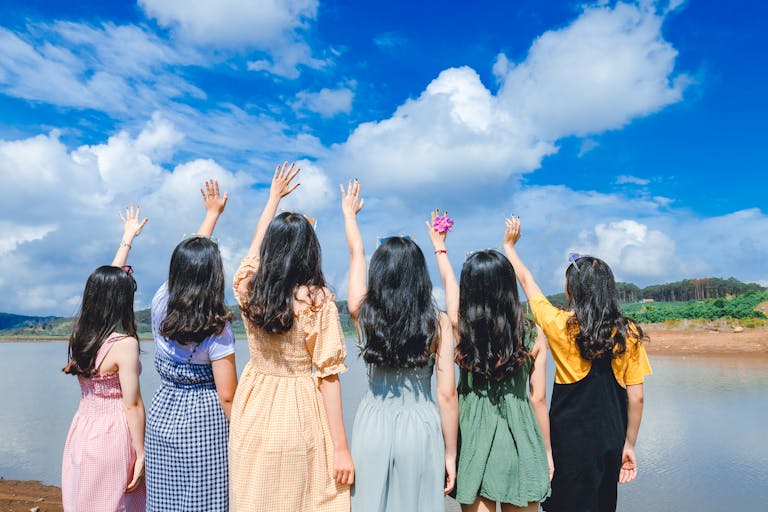 Group of young women with long hair raising hands by a lake under a clear blue sky.