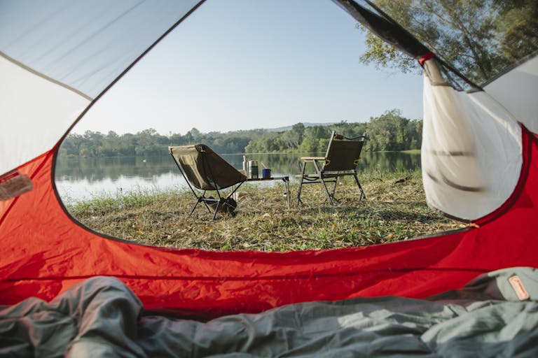 Relaxing campsite view from a tent by a peaceful lake with chairs under clear skies.