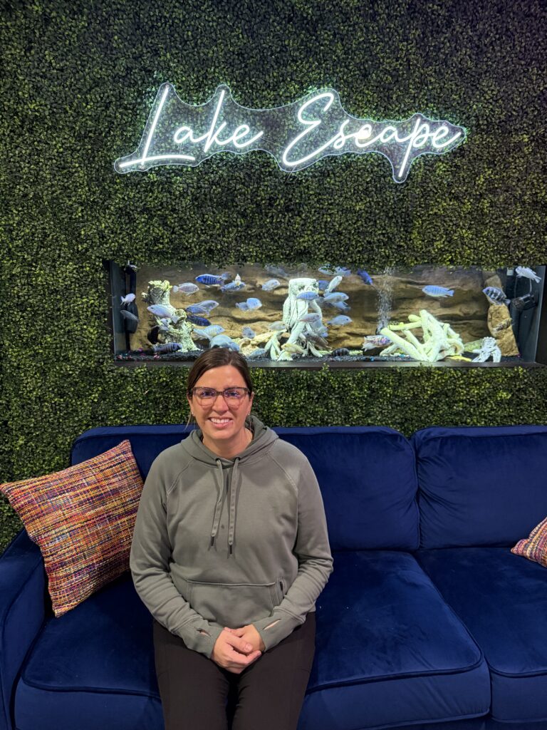 Photo of woman sitting on a navy velvet couch in front of a neon Lake Escape sign hanging on a green plant wall with a fish tank in the background