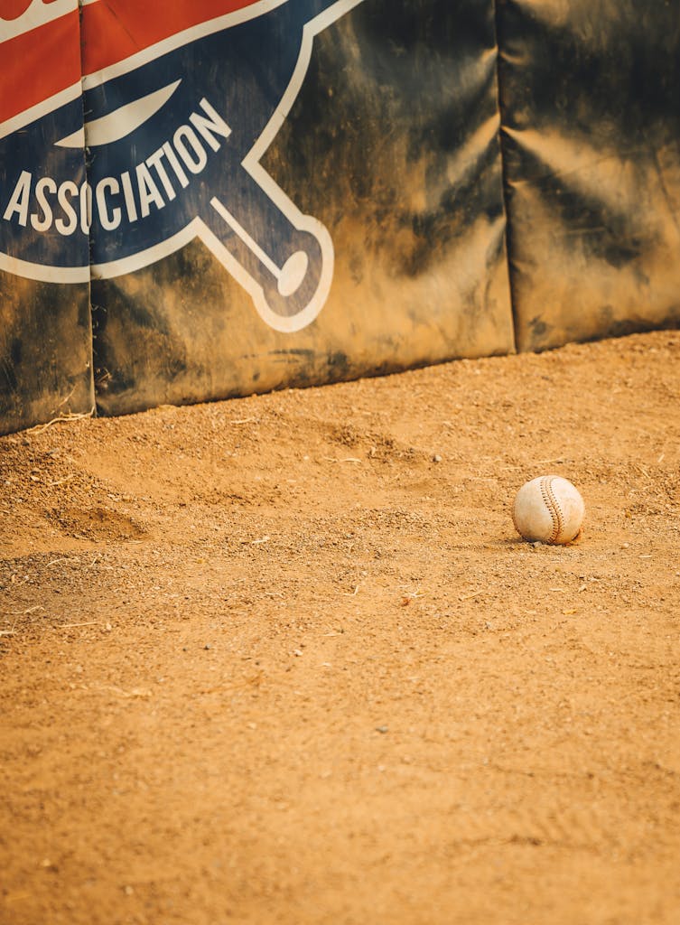 A baseball rests on a sandy field in front of a sports association banner, capturing a moment in outdoor sports.