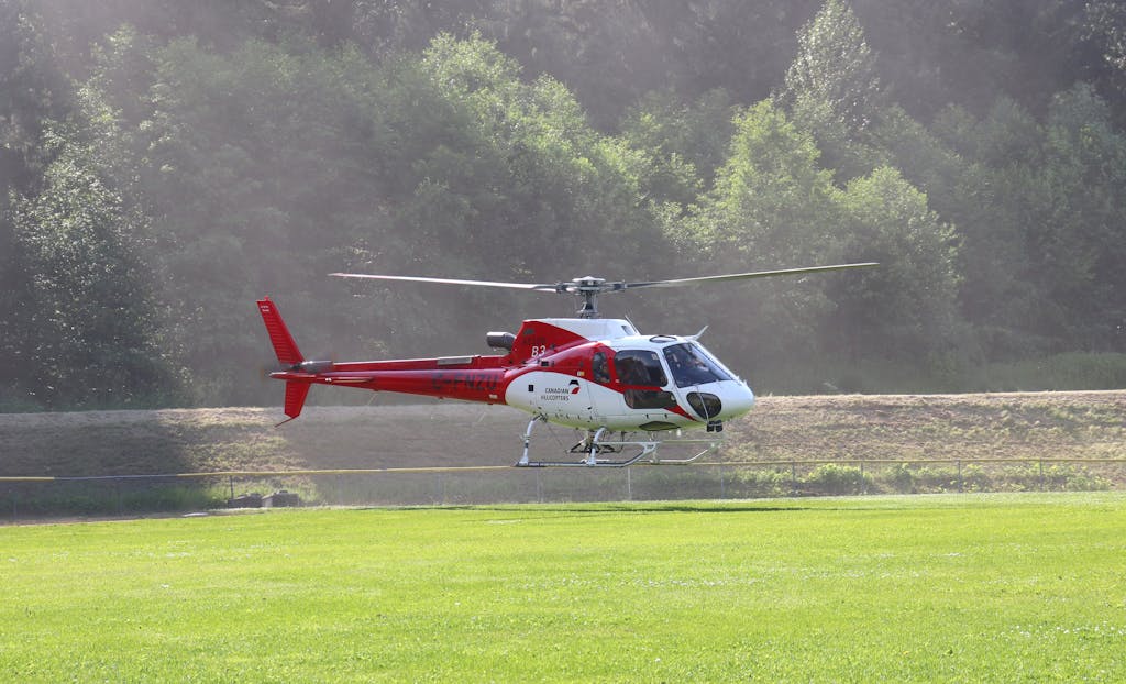 Aerial view of a red and white helicopter flying above a green field against a forest backdrop.