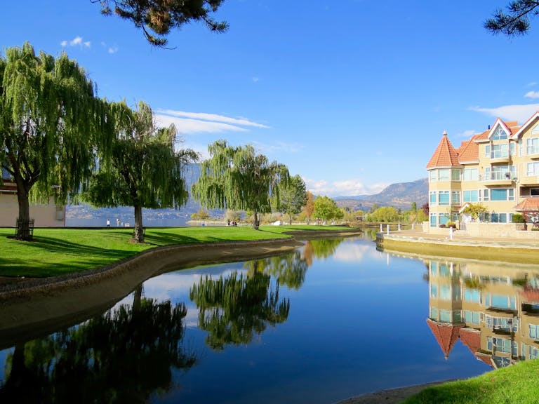 Beautiful lakefront view with reflections, trees, and a luxury building in a sunny setting.