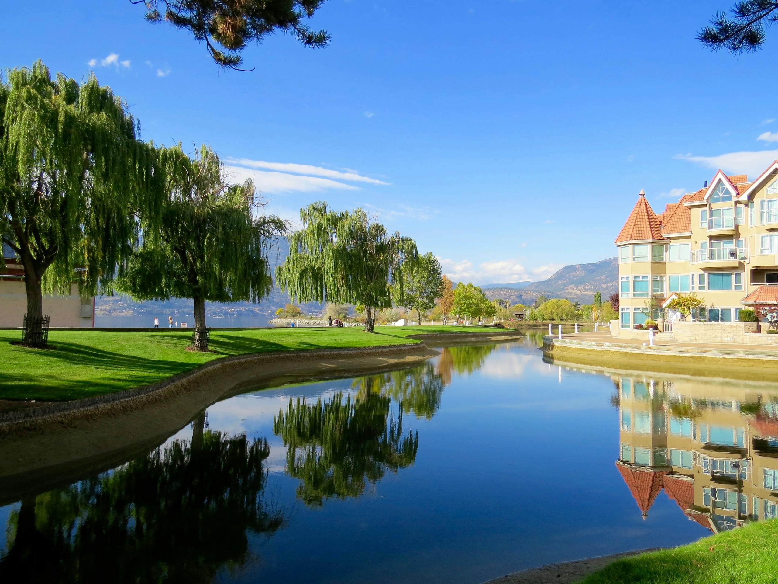 Beautiful lakefront view with reflections, trees, and a luxury building in a sunny setting.