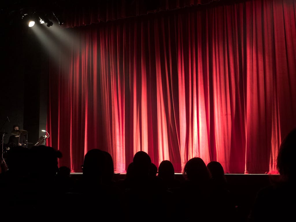 Dimly lit theater stage with red curtains and audience silhouettes under spotlights.