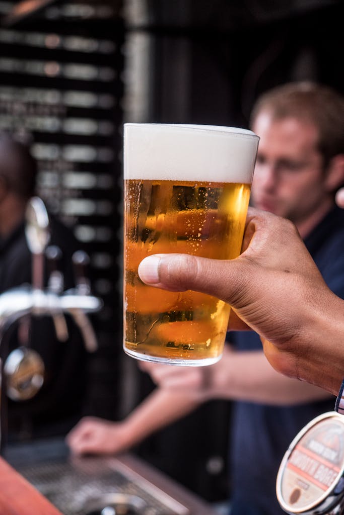 Close-up of a hand holding a foamy beer glass with blurred bar background.